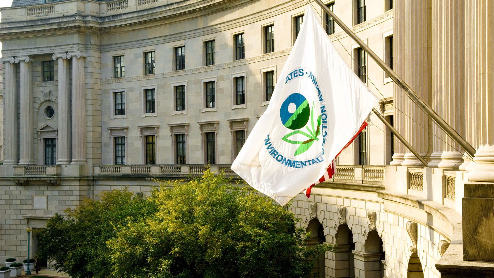 A white flag carrying the seal of the United States Environmental Protection Agency hangs from an angled pole on the front of the William Jefferson Clinton Federal Building in Washington, the agency's headquarters. An American flag hangs partly hidden behind it. The pale stone facade curves away in rows of tall windows above a ground floor arcade, with columns at the right and a tree in full leaf below in warm late light.