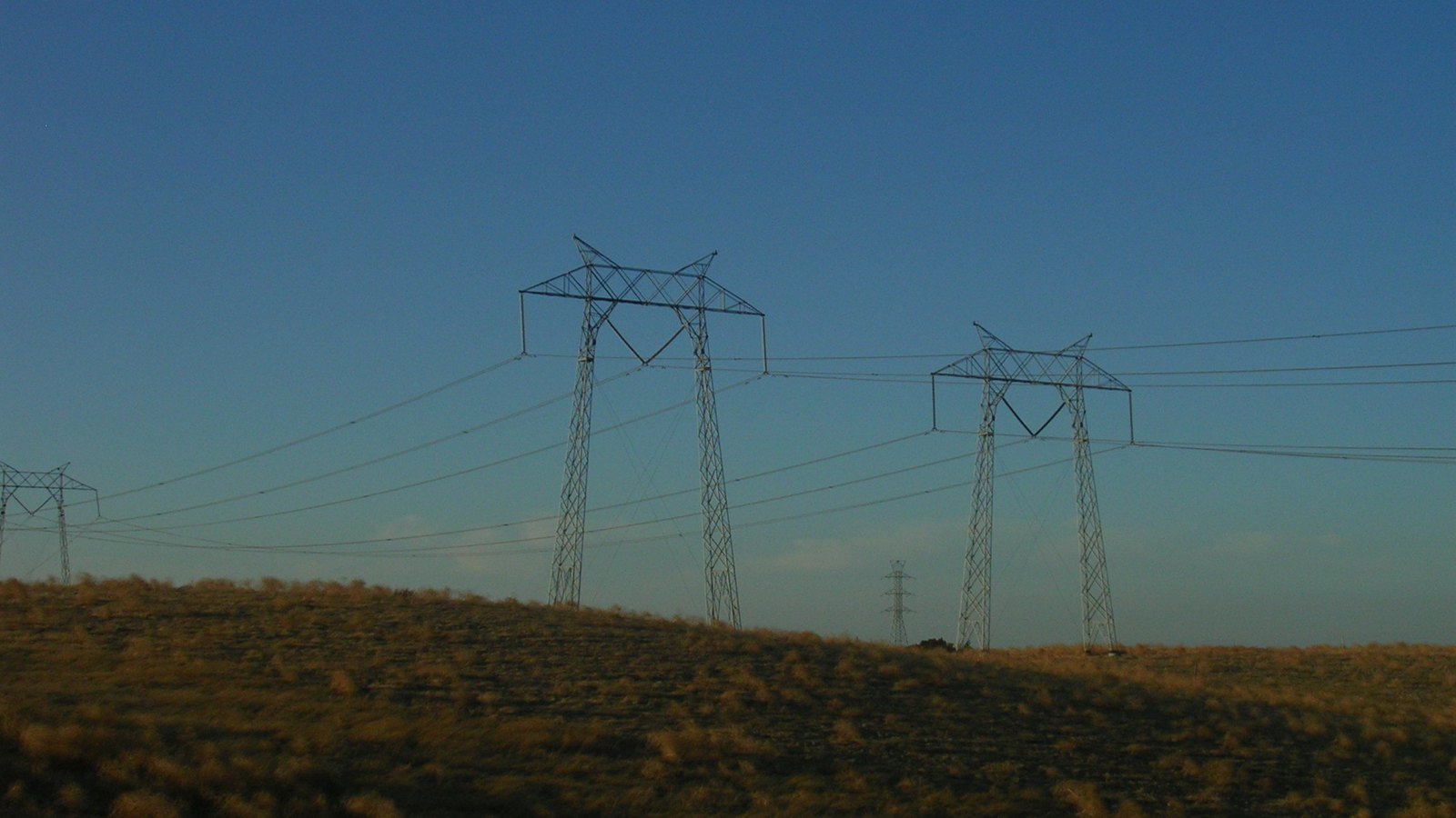 Two lattice steel transmission towers carrying high-voltage lines over a dry grassy hill against a clear blue evening sky, with smaller towers in the distance.
