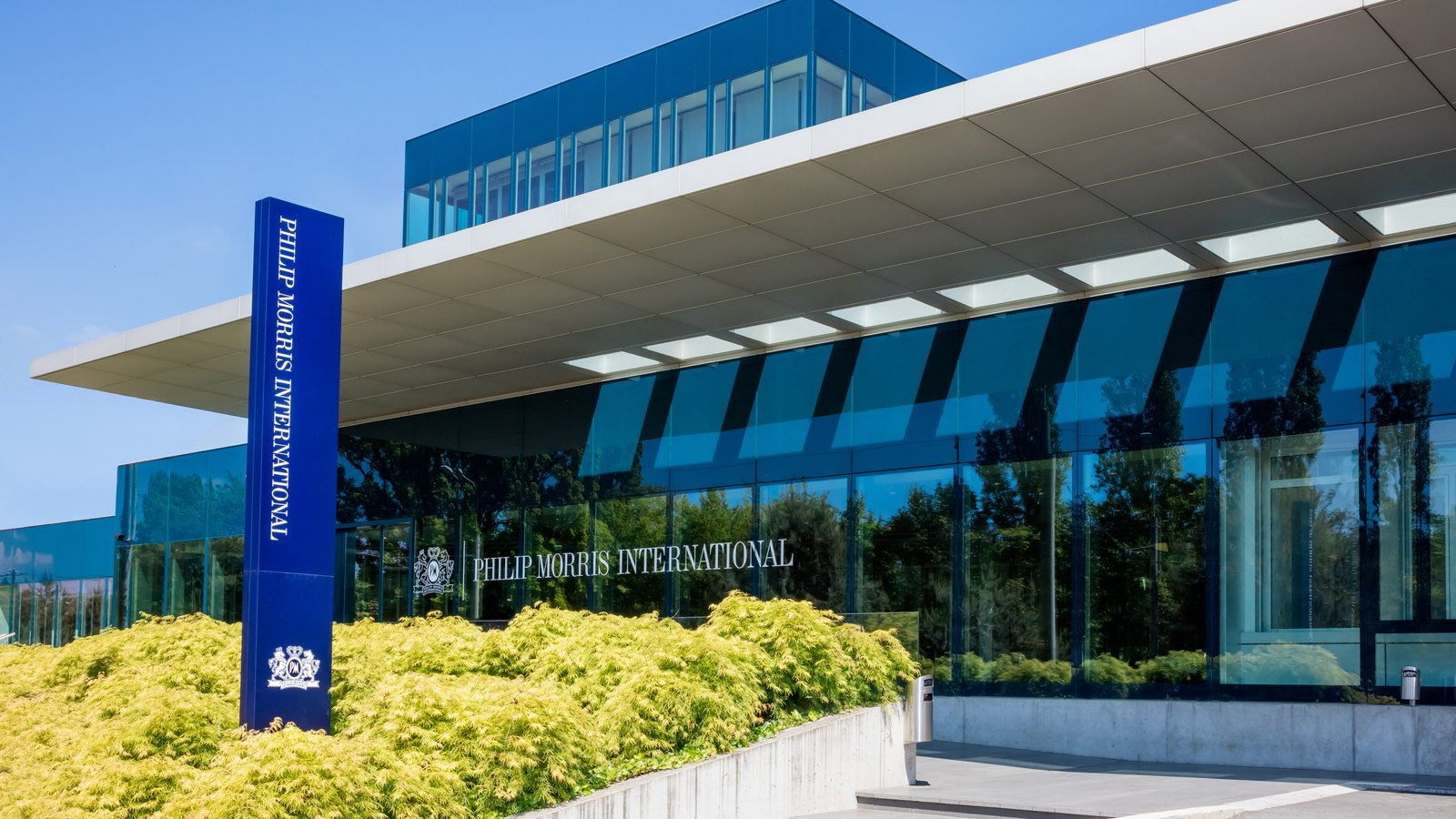 The glass and steel entrance of a Philip Morris International office building in Lausanne, Switzerland, with the company name on a blue pylon and on the facade above yellow-green shrubs.