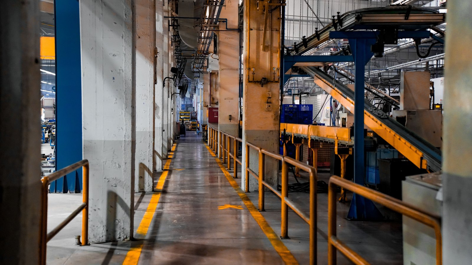 A yellow marked walkway runs down the middle of a working factory, between concrete columns on one side and machinery, guard rails and an overhead conveyor on the other. No brand mark, sign or readable word is legible in the frame, and no person is close enough to be identified.