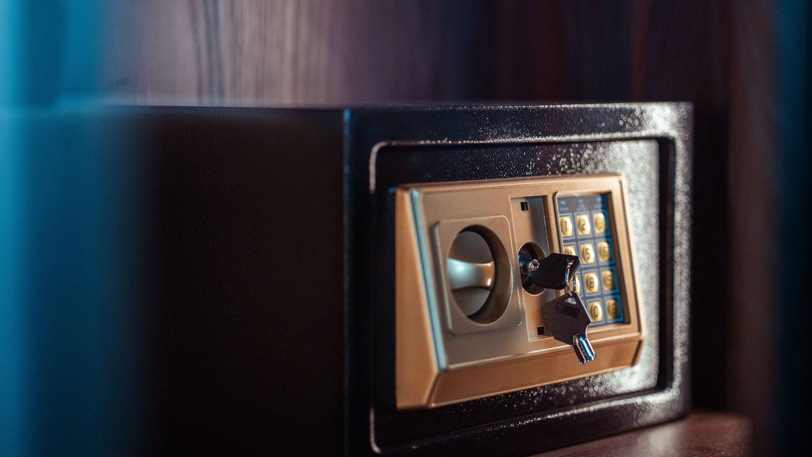A small black room safe with a gold keypad and dial sits on a dark wood shelf, a key left in the lock beneath the keypad. No person, room number, signage or lettering of any kind is visible. Stock photo, not a photograph of this event.