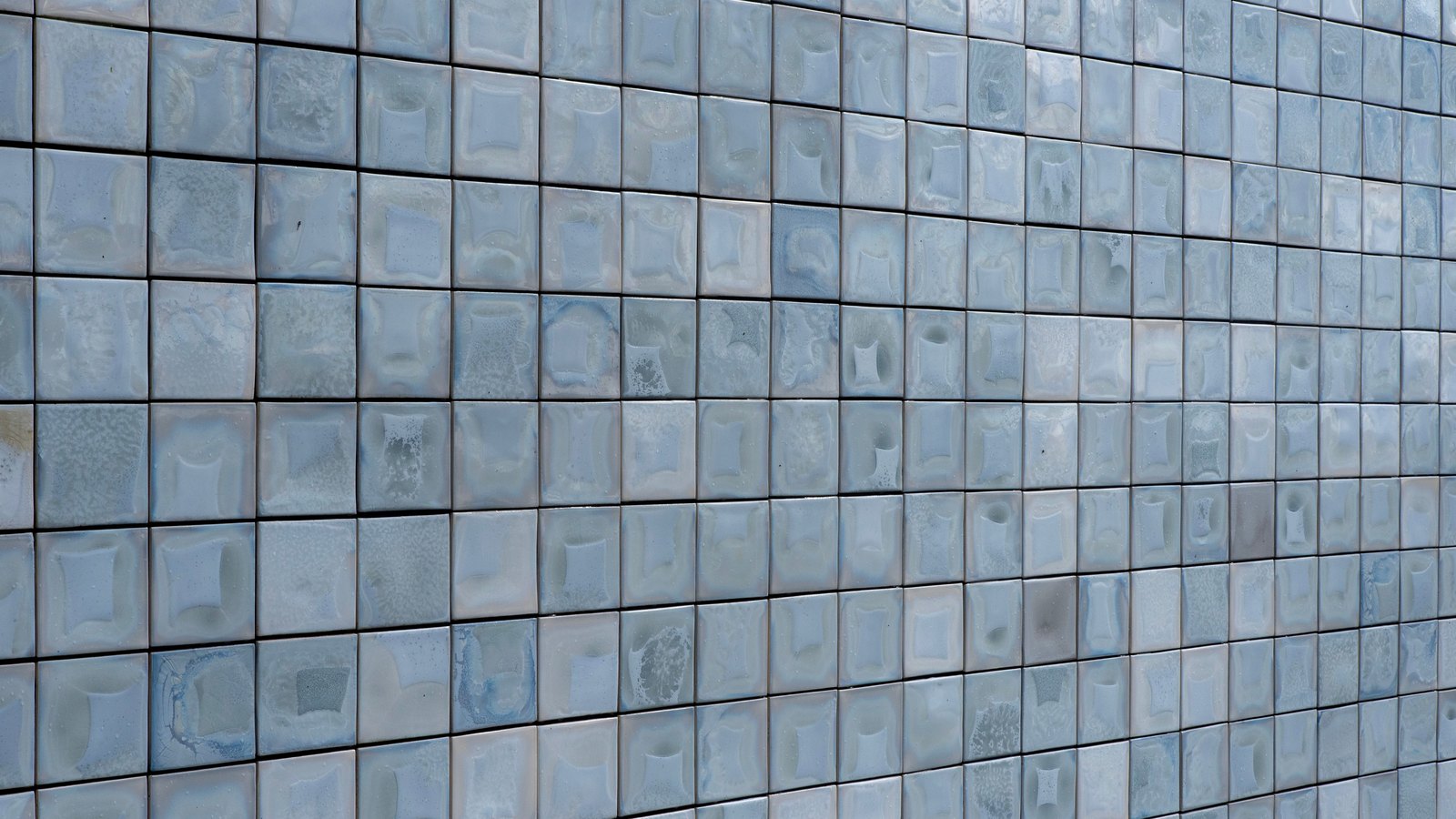 A wall of small pale blue grey glazed ceramic tiles filling the whole frame, hundreds of squares in even rows receding slightly to the right, each one lightly domed and separated by a thin dark joint. No text, brand mark, person, fixture or sky is in view.