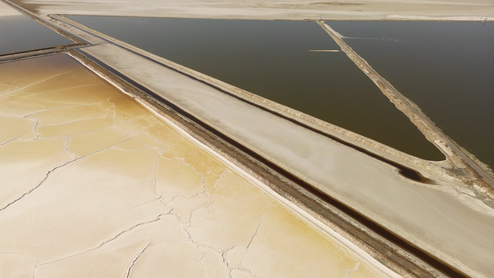An aerial view of industrial salt evaporation ponds, a straight earth causeway running diagonally between a wide pond of standing brine and a pale crystallising pan whose dried crust has broken into a web of cracks, with no buildings, vehicles, people or lettering in the frame.