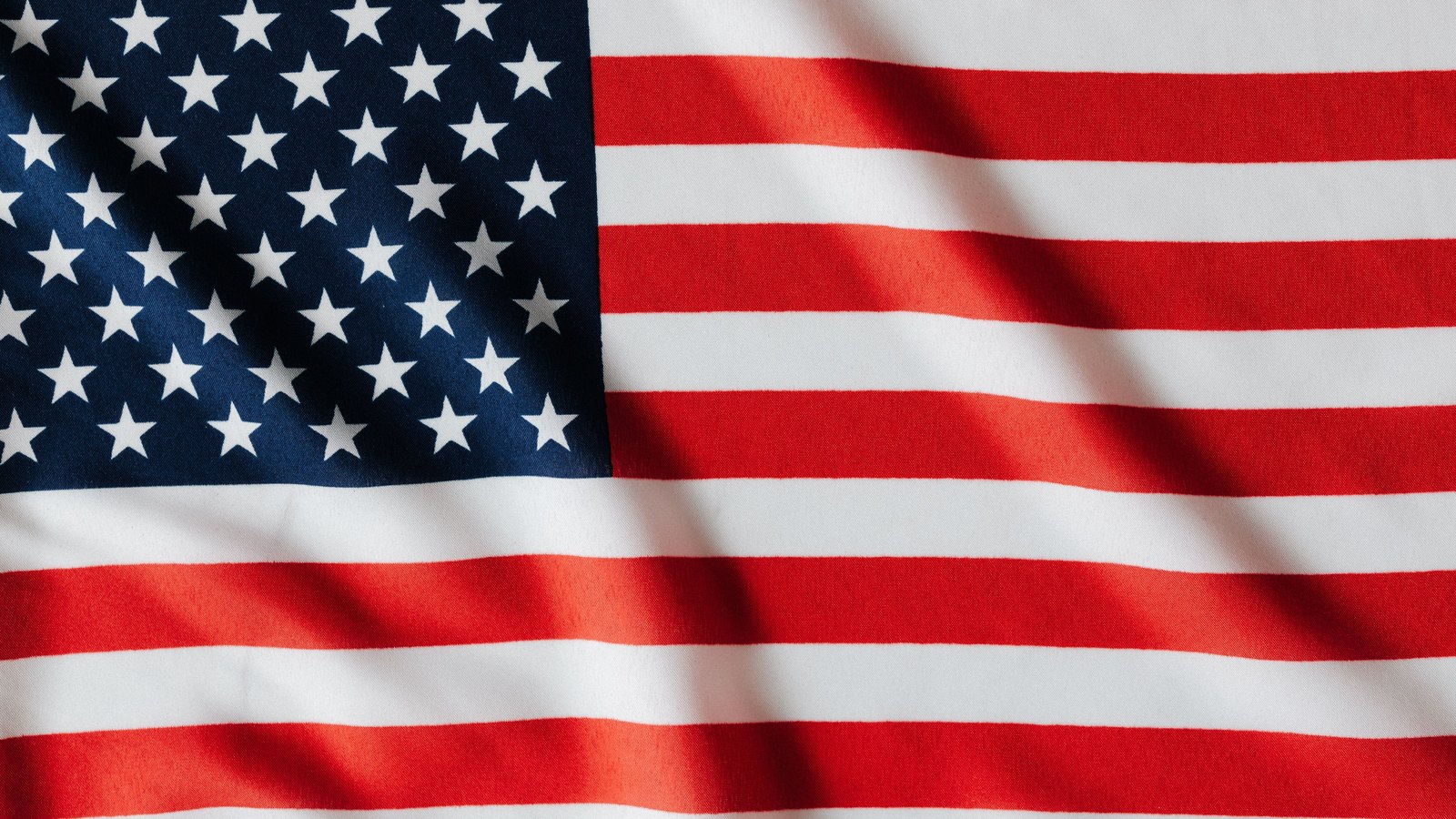 A close up of the flag of the United States filling the whole frame, shot flat against rippled cloth. The blue canton with its white stars sits at the upper left and the red and white stripes run to the right edge. Soft diagonal shadows fall across the fabric where it folds. No pole, building, person, signage or text is in view.
