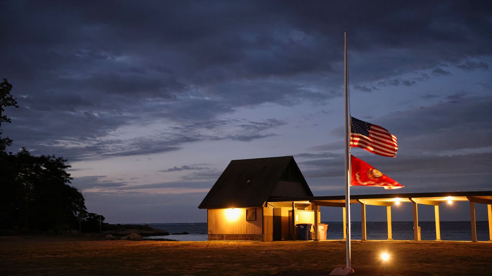A United States flag flying at half-staff on a white pole at dusk, with a Marine Corps flag below it, both lit from below. A small shingled shelter and a lit open pavilion stand behind on a grass foreshore, with dark water and heavy blue cloud beyond. No lettering or signage is legible.