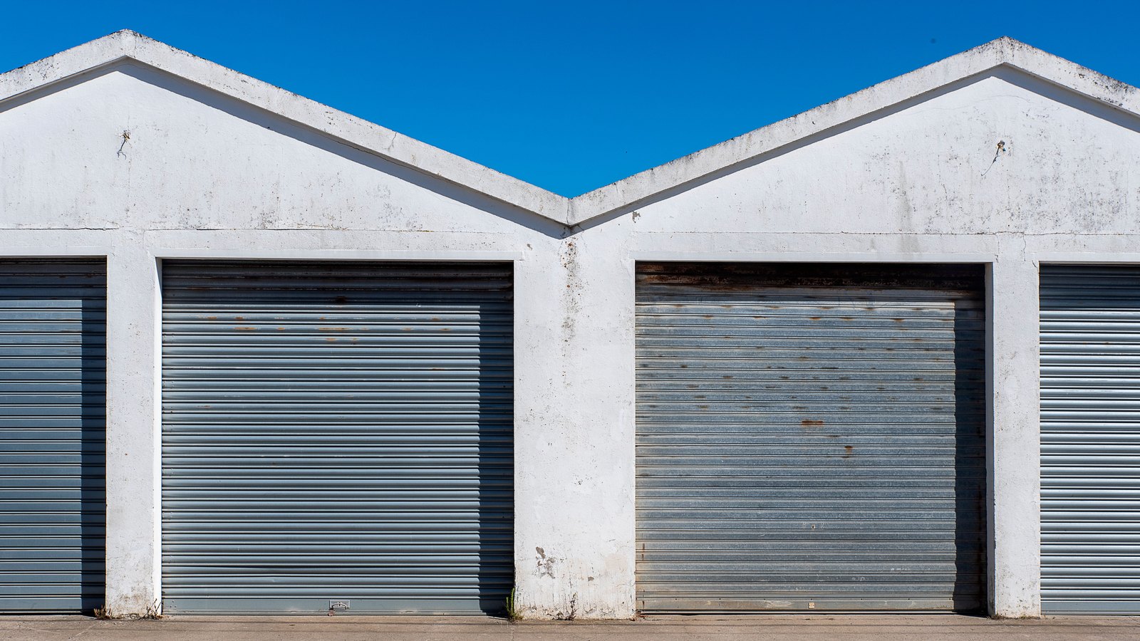 A row of white self-storage units with grey metal roll-up doors under a clear blue sky.