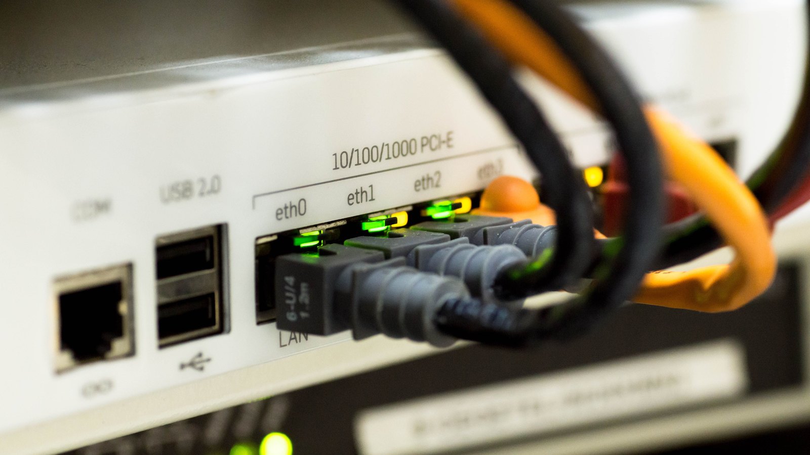 A close up of the rear panel of a rack-mounted network appliance, with grey ethernet cables plugged into ports labelled eth0, eth1 and eth2 above a row of lit green and amber link lights, two USB sockets and an unused socket to the left, and black and orange cables curving across the top of the frame.