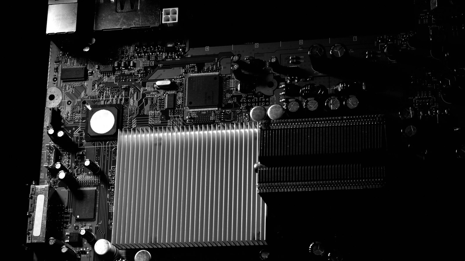 A low key, almost monochrome close photograph of a computer motherboard, with a brightly lit finned aluminium heatsink at the centre, empty memory slots to the right and rows of small components fading into black, carrying no brand name.