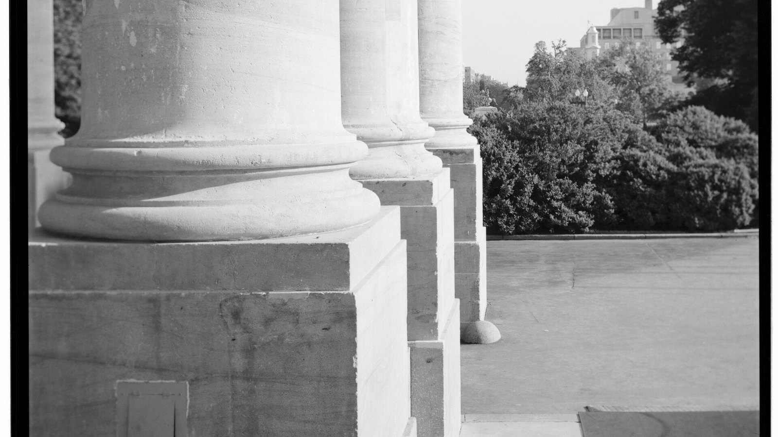 A black and white photograph of three stone column bases in a receding row on the north portico of the White House, each standing on a stepped stone plinth, with a paved terrace to the right and trees and a distant building beyond. No person or lettering is visible.