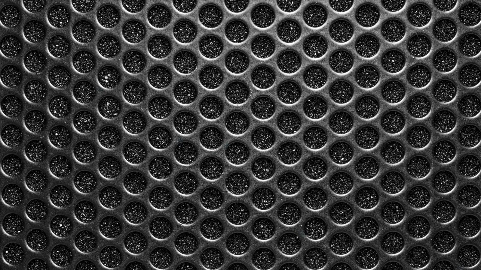 A close photograph of a dark metal speaker grille, filling the whole frame with rows of identical round holes pressed into the panel, each ring catching a thin highlight along its upper edge and each opening showing the coarse black mesh behind it, with the light falling away towards the lower corners.
