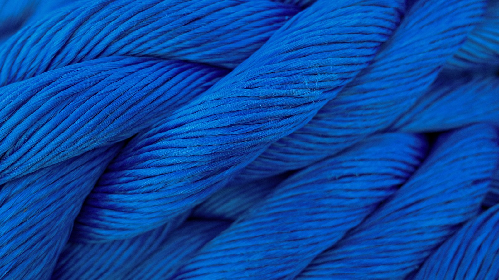 A close photograph of thick blue rope filling the entire frame, several lengths lying side by side and running diagonally across it, each one twisted from three strands so that fine parallel fibres catch the light along every turn. A few loose fibre ends stand up from the surface. No text, brand mark, person, place or object edge is in view.
