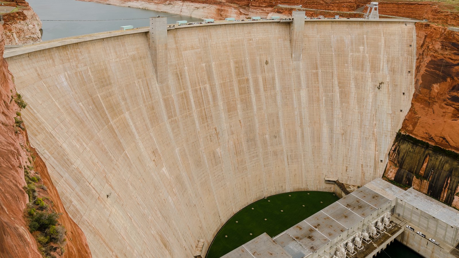The downstream face of Glen Canyon Dam, a pale concrete arch filling the frame between red sandstone canyon walls, with the powerhouse and switchyard at its foot and a strip of Lake Powell visible beyond the crest.