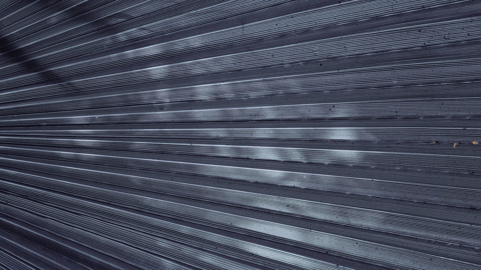 Row after row of narrow parallel ribs in a cool blue grey metal surface, receding across the whole frame, with soft dappled light falling on them. No text, brand mark, person, horizon or building is in view.