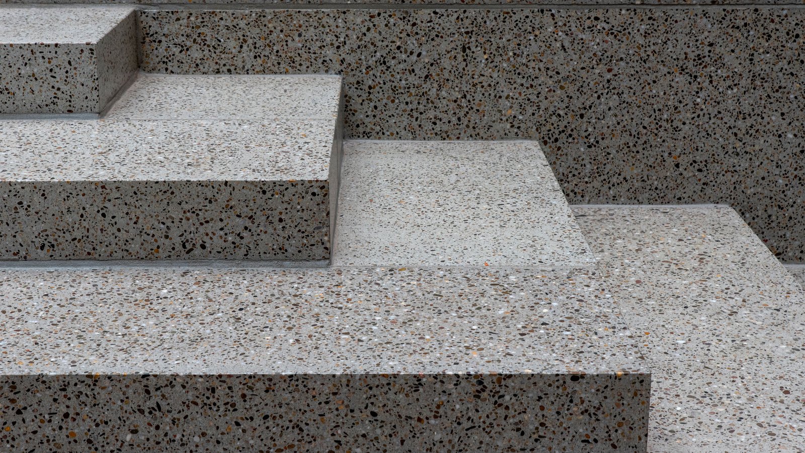 Polished terrazzo steps photographed straight on, pale grey slabs flecked with brown, black and orange aggregate, the treads and risers stepping across the frame in flat geometric bands, with no signage, handrail or person in view.