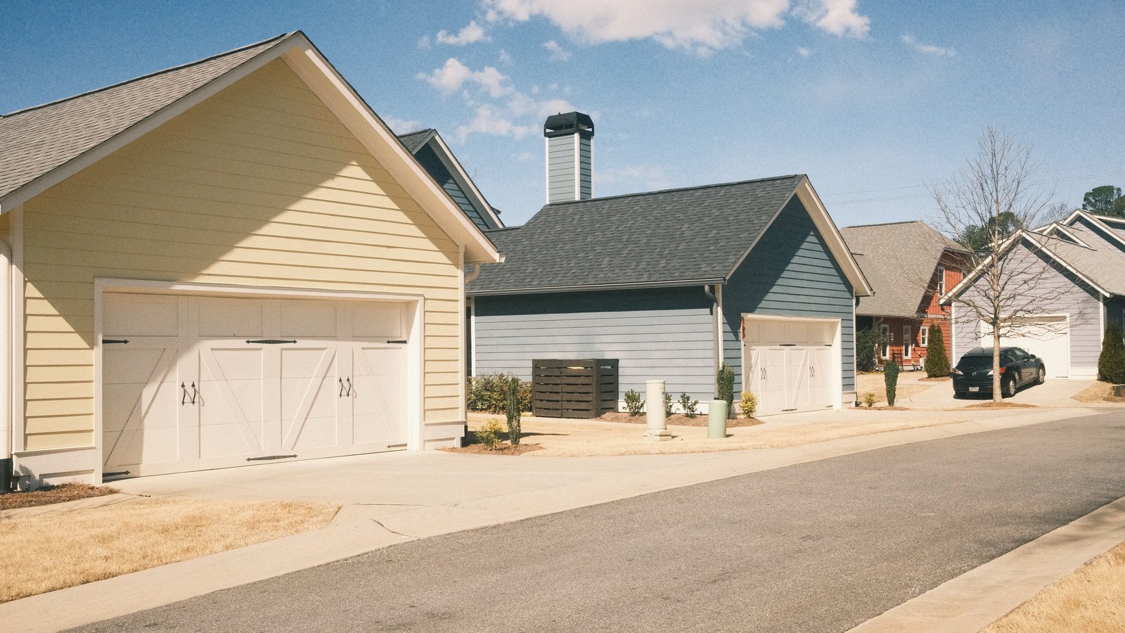 A row of newly built American suburban houses in cream and pale blue lap siding, each with a white carriage style garage door and a concrete driveway, along an empty paved street under a clear sky.