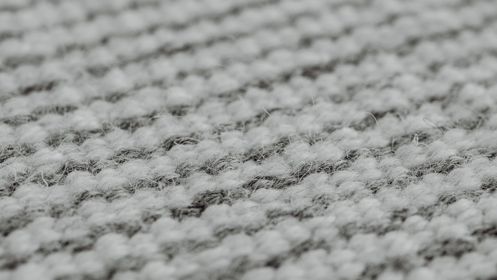 Coarse grey wool cloth photographed at a low angle so the weave runs away out of focus. Rows of pale nubby loops alternate with darker, more tightly spun rows, and a few loose fibres stand up against the light. No lettering, edge or object appears anywhere in the frame.