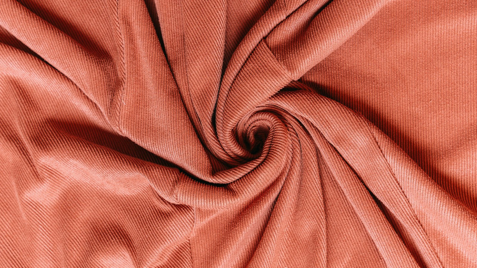 A length of ribbed corduroy cloth in a warm terracotta pink, twisted into a spiral at the centre of the frame so the fine parallel ridges curve around it. The cloth fills the whole picture and carries no lettering or marking other than its own ribs.