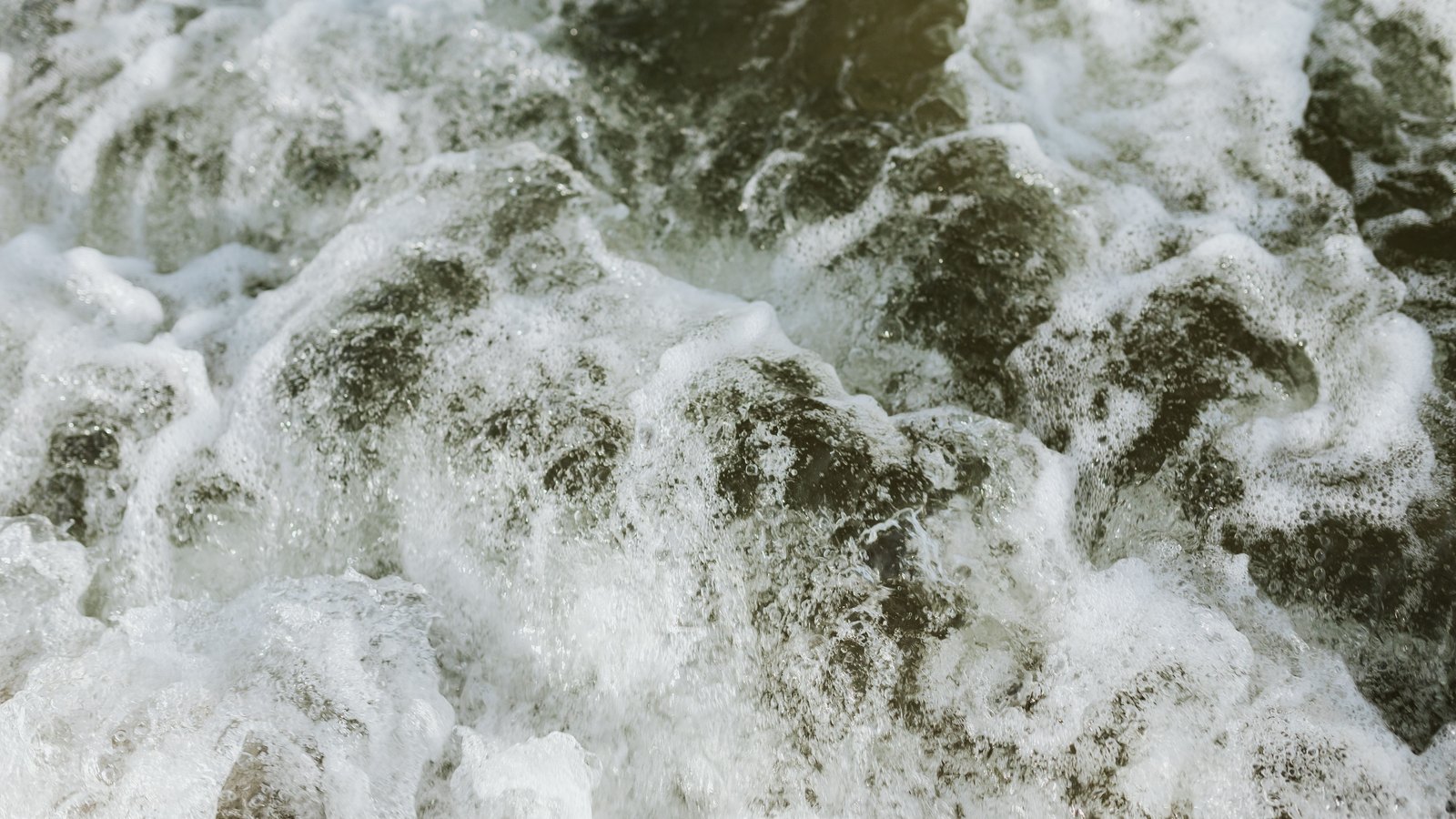 A close overhead crop of turbulent surf, ridges of white foam breaking across dark green water, filling the frame with no horizon, shoreline, people or text in view.