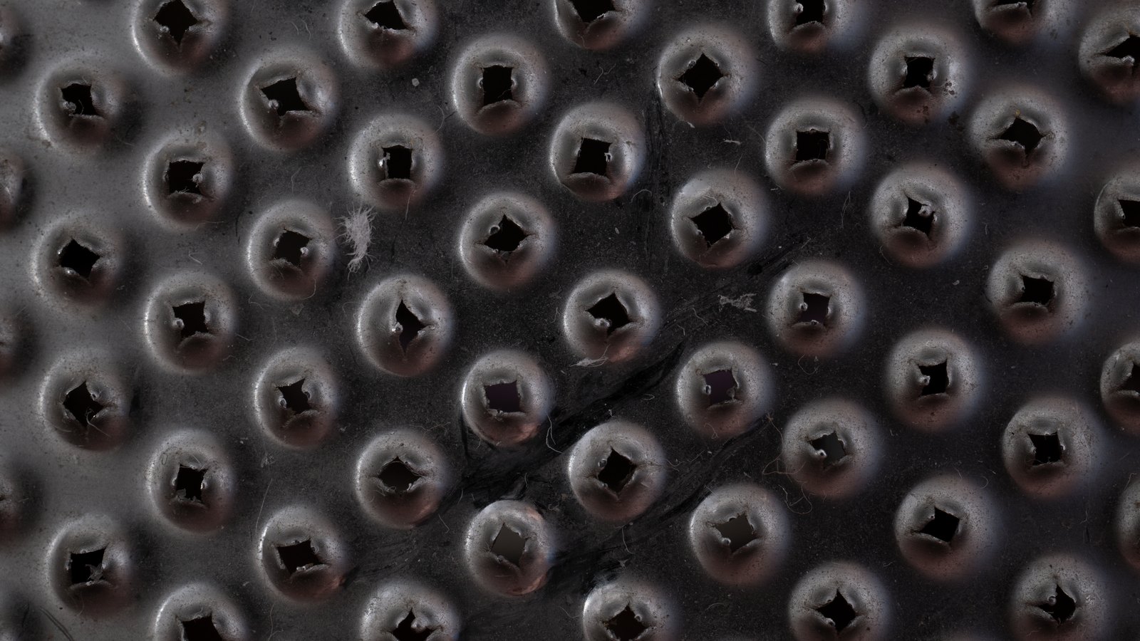 A close photograph of a dark metal sheet punched all over with raised circular bosses, each pierced by a small square hole, the surface scuffed and lit from one side, filling the whole frame with no text, label or background visible.