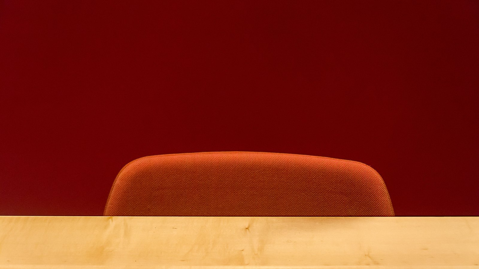 An empty upholstered chair in muted orange, its back just visible above the pale wooden edge of a desk, against a plain deep red wall. Nothing else is in the frame.