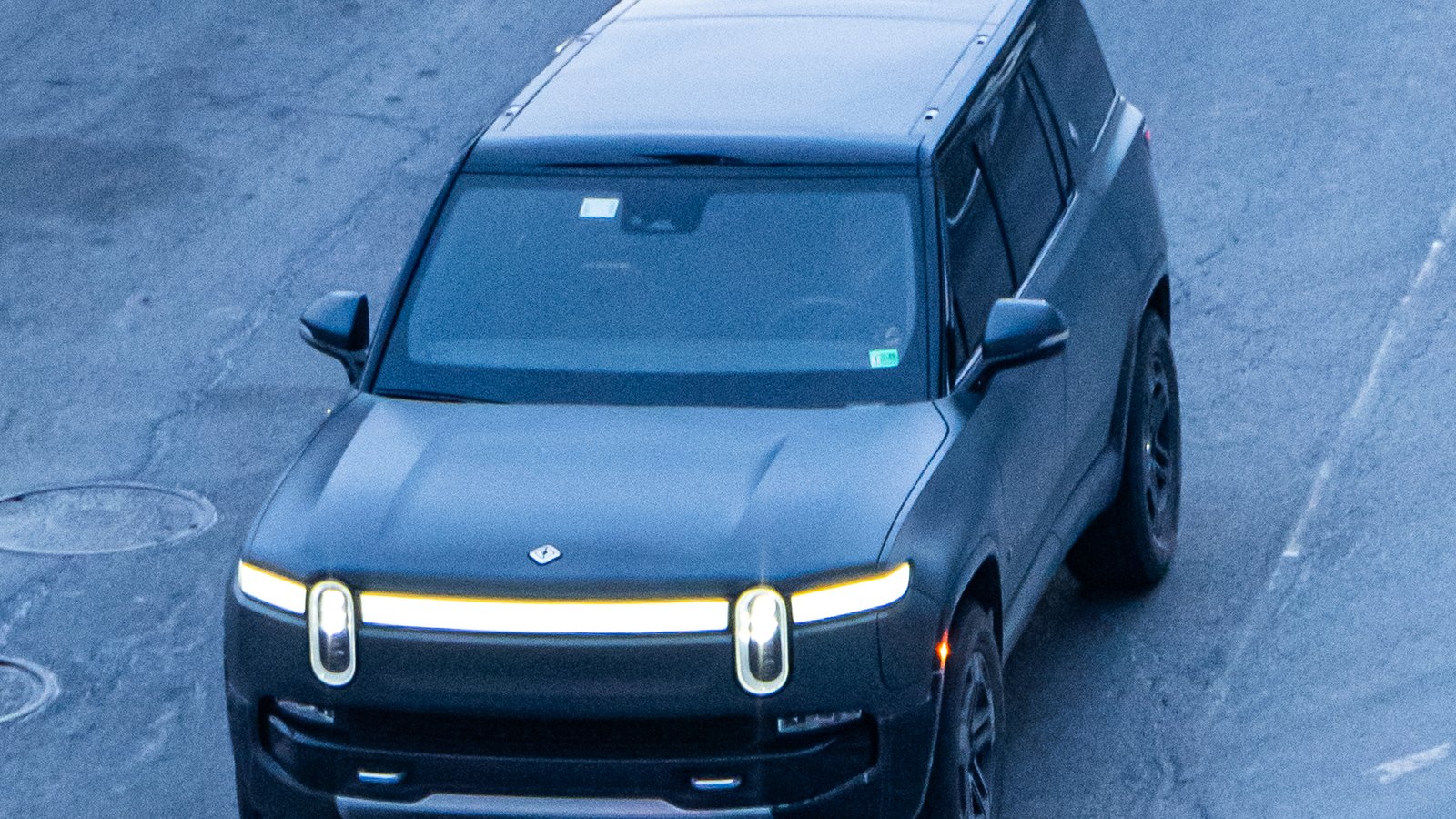 A dark Rivian R1S drives across a marked roadway seen from above
