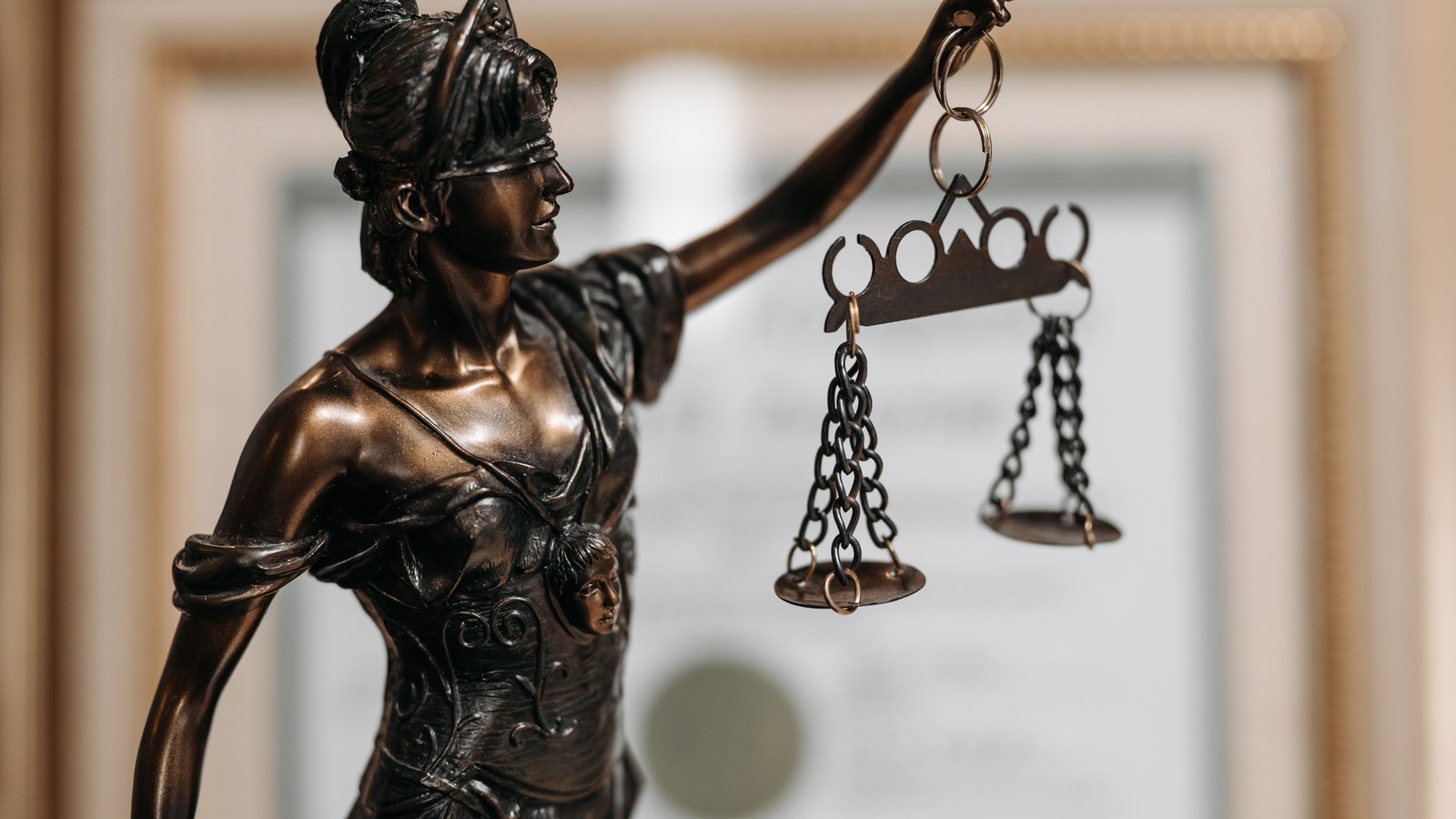 A close photograph of a bronze Lady Justice statue holding balance scales against a softly blurred office background, with no people, place names or readable text visible, not a photograph of this event.