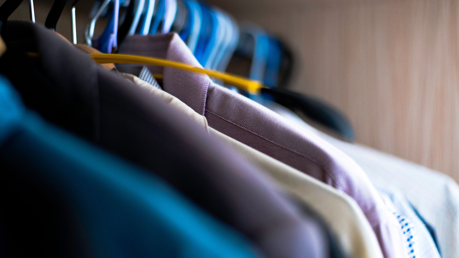 A close view along a rail of shirts on hangers, shoulders overlapping in teal, brown, mauve, cream and pale grey, with a row of blue and white striped shirts out of focus behind them. No labels or branding are visible.