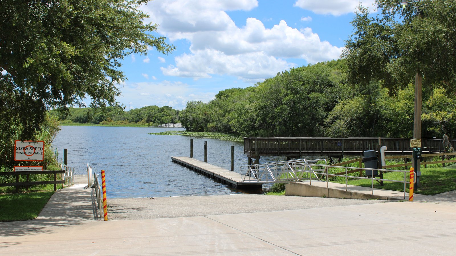 A concrete boat ramp sloping into a calm tree lined river, with a floating dock and pilings to the left, a raised wooden boardwalk with a handrail running along the far bank, live oaks and lily pads around the water and cumulus cloud overhead, and a small generic slow speed minimum wake waterway sign at the left edge. No people, no place name, no agency markings and no number plates are visible. A generic illustration, not a photograph of this event.