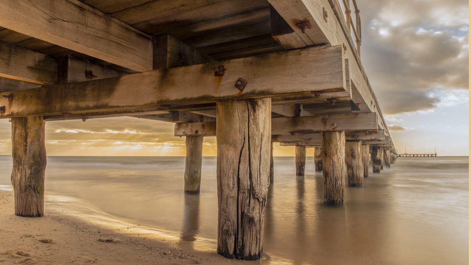 The underside of a timber pier seen from a sandy shore, its plank deck and cross beams resting on weathered round wooden pilings that stand in shallow, still water under a hazy gold sunset sky. No person, vehicle, building, signage or lettering of any kind is visible in the frame. Stock photograph, not a photograph of this event.