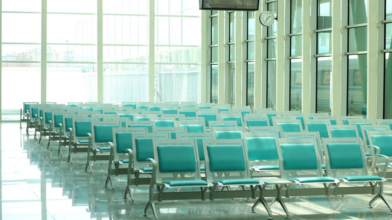 Rows of empty turquoise vinyl seats on white metal frames filling a bright airport departure lounge, with a floor to ceiling glass wall, a wall clock and an overhead flight information screen whose text is not legible. No person, airline marking or place name is in view. Illustrative only, not a photograph of this event.