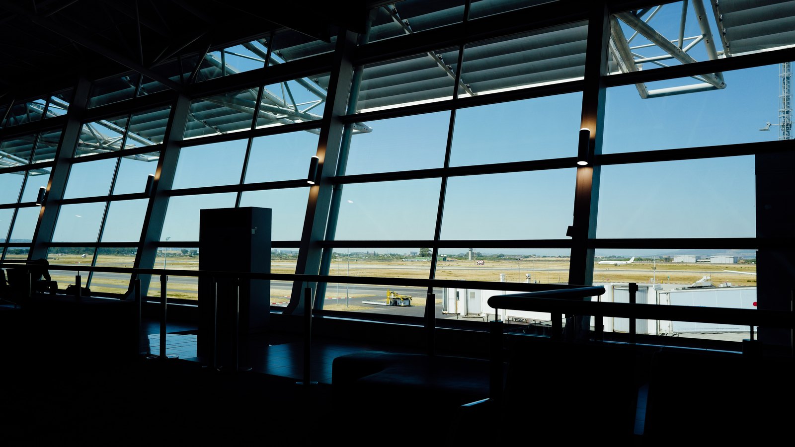 Interior of an airport terminal in silhouette, looking through a tall window wall onto an empty apron and runway under a clear blue sky, with dry tan grass beside the taxiway, a jet bridge at the right and a small yellow ground vehicle in the distance. No people, no signage and no aircraft markings are visible. Illustrative only, not a photograph of this event.