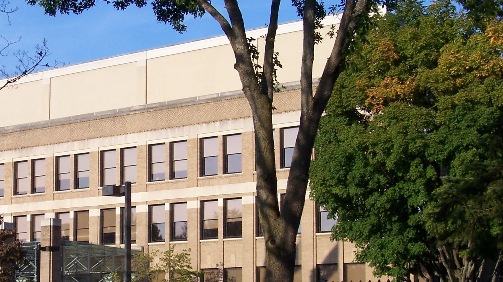 The pale brick facade of the Pratt and Whitney headquarters building in East Hartford, Connecticut, with rows of windows and a patterned frieze behind a large tree in early autumn leaf under a blue sky.