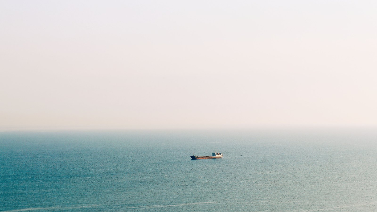 A distant cargo ship crosses calm water on the Black Sea below a pale sky.