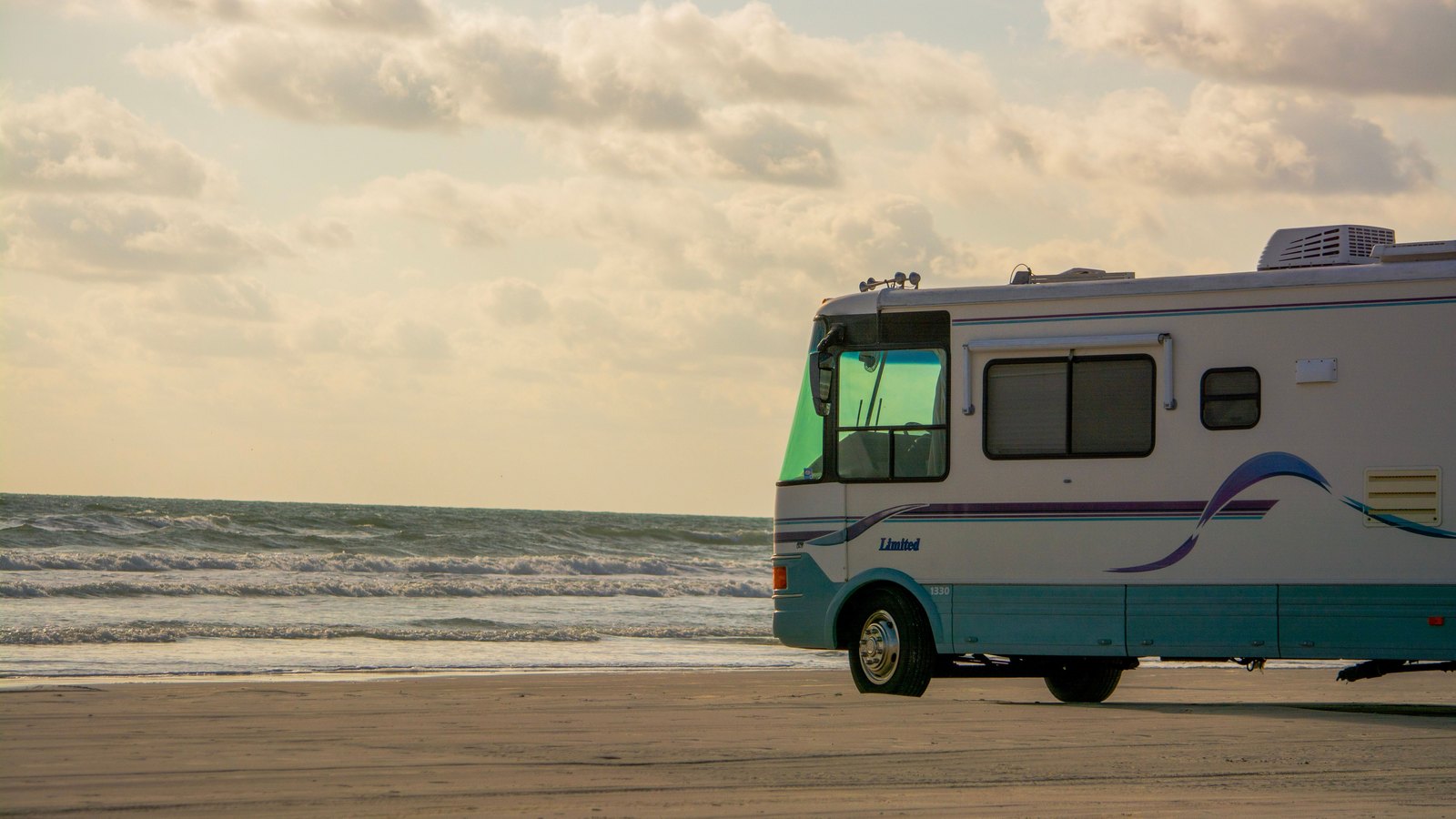 The front half of a large white American motorhome with teal and purple side striping, parked on wet sand at the edge of a beach with low surf and a hazy, cloud-streaked sky behind it. No person is in frame and no plate or business name is legible. Stock photo, not a photograph of this event.