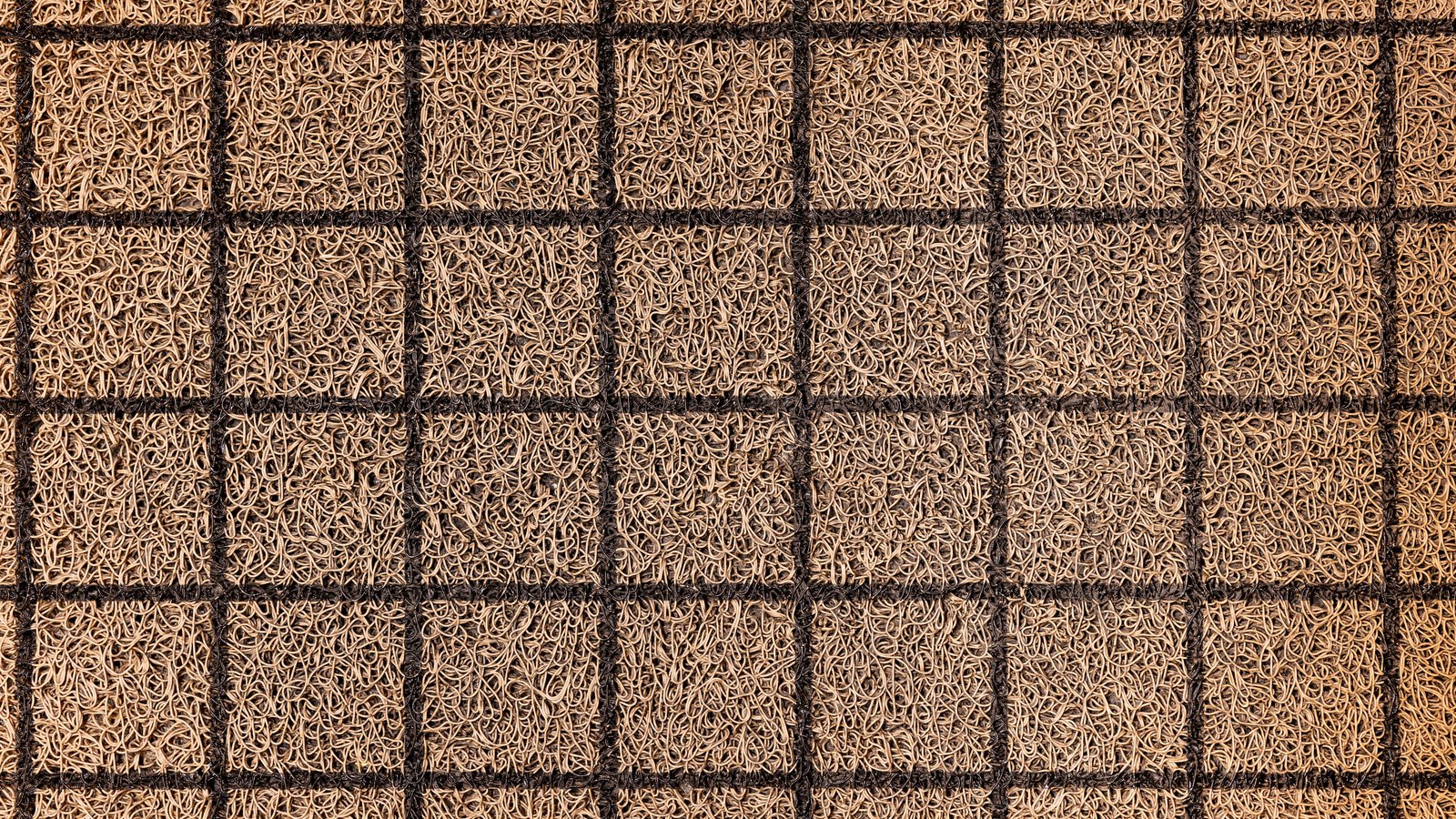 A close photograph of a woven mat filling the whole frame, tan looped fibre packed into a grid of squares separated by dark ribs and running off every edge. It is used as a generic illustration and is not a photograph of either hotel. No text, brand mark, person or place is in view.