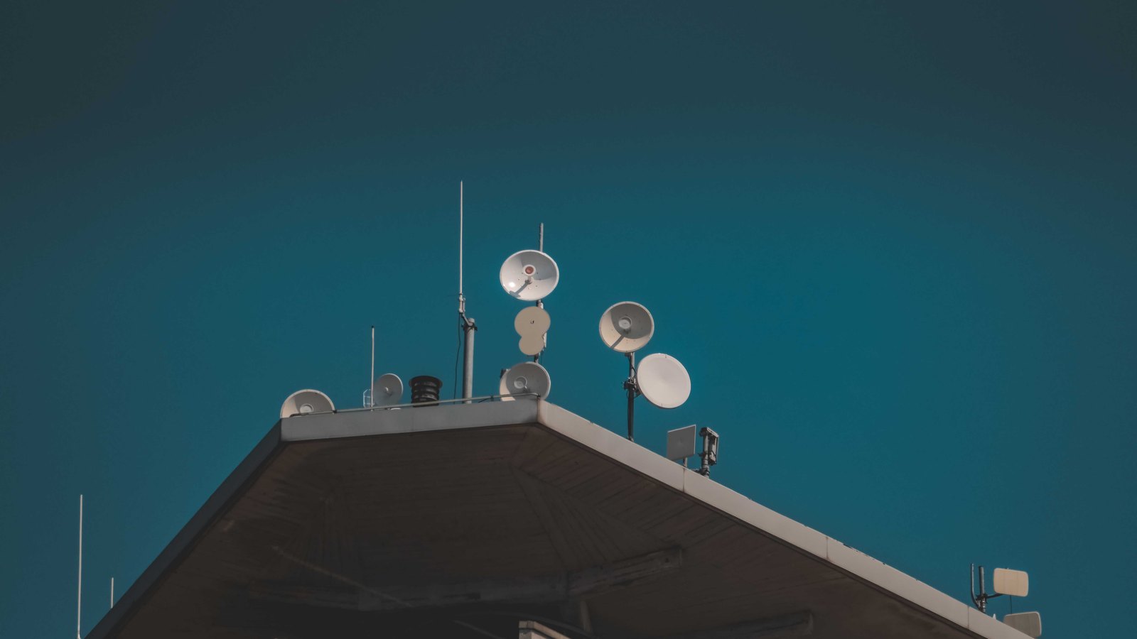 A cluster of white microwave dishes and whip antennas on the ridge of a flat roof, seen from below against an empty dark blue sky.