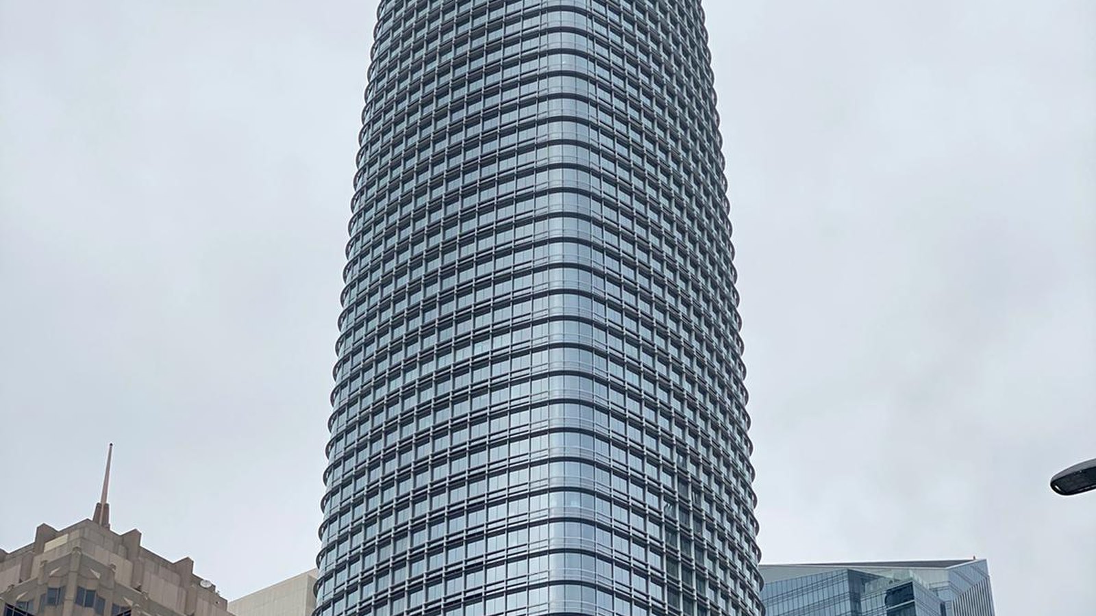 Salesforce Tower in San Francisco seen from the street below, a tapering glass tower with rounded corners and closely spaced horizontal bands of windows rising into a flat white overcast sky, with the stepped stone crown of an older building at the lower left and a low glass block at the lower right.