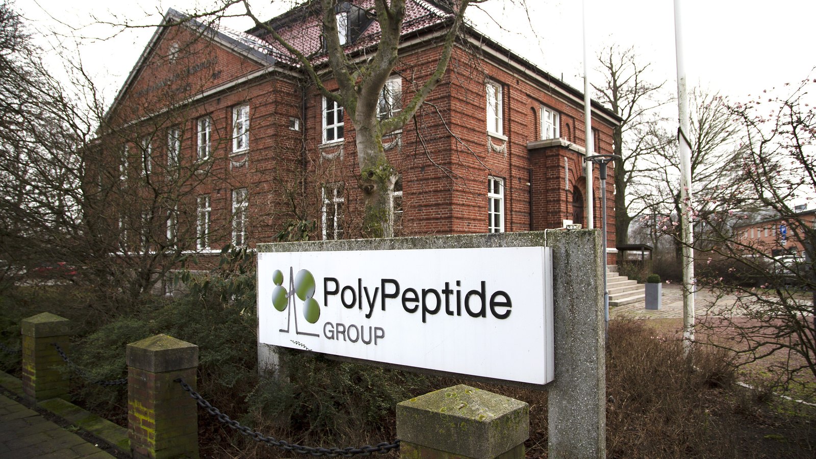 A white sign on a low concrete plinth reading PolyPeptide Group, the company logo of green circles beside the wordmark, standing at the entrance to a two storey red brick building with a tiled roof and white framed windows. Bare winter trees, a clipped hedge and two flagpoles surround it under a flat white sky. Photographed at the company site in Malmo in January 2020.