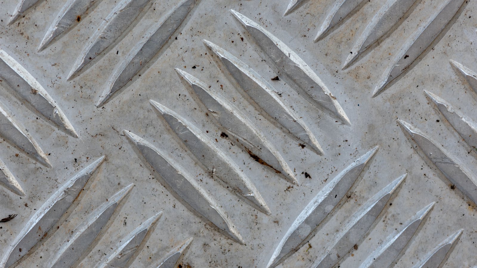 A close overhead macro of bright aluminium diamond tread plate, its raised lens-shaped ridges running in alternating diagonal rows across the whole frame, the flat metal between them scuffed and lightly speckled.