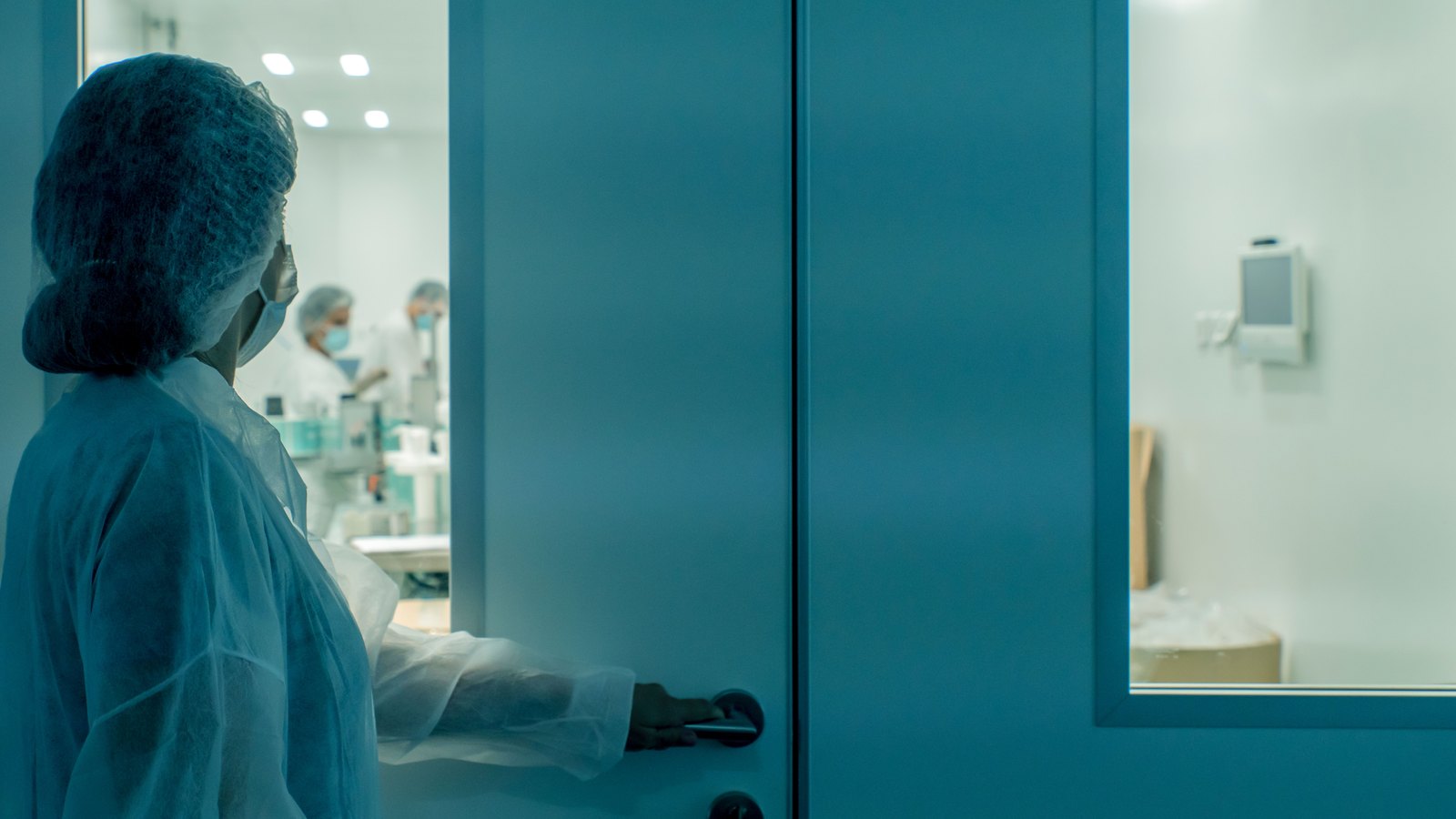 A masked laboratory worker in protective clothing enters a sterile cleanroom.