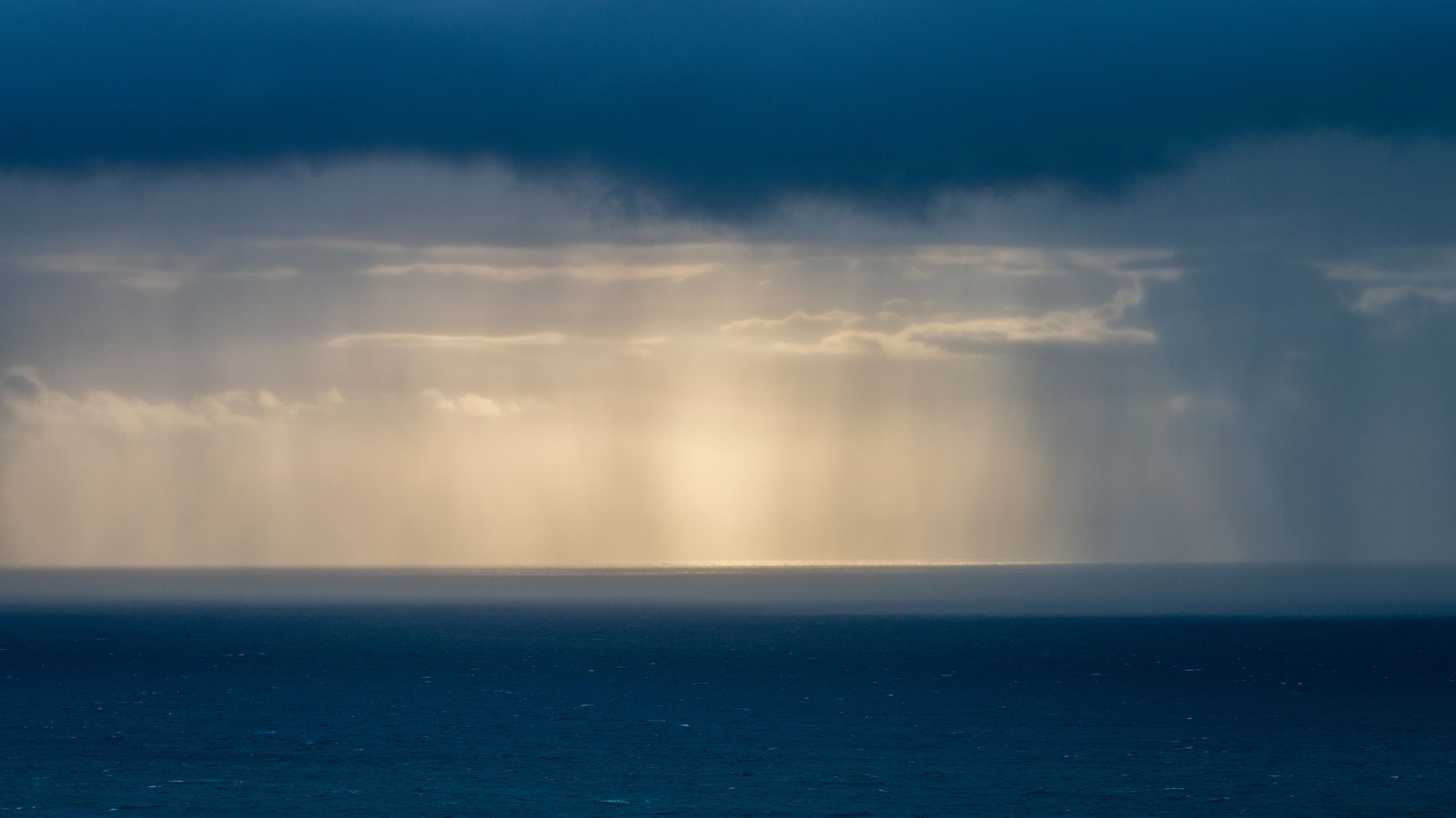 A dark blue storm cloud layer spread across the top of the frame above open sea, with a broad shaft of rain falling from it and a band of pale gold light breaking through low on the horizon. No shoreline, bridge, road, vehicle, person, structure or legible text is in view, and no location can be identified from it. A plain illustration of a storm over water, not a photograph of the Sanibel Causeway or of this fire.