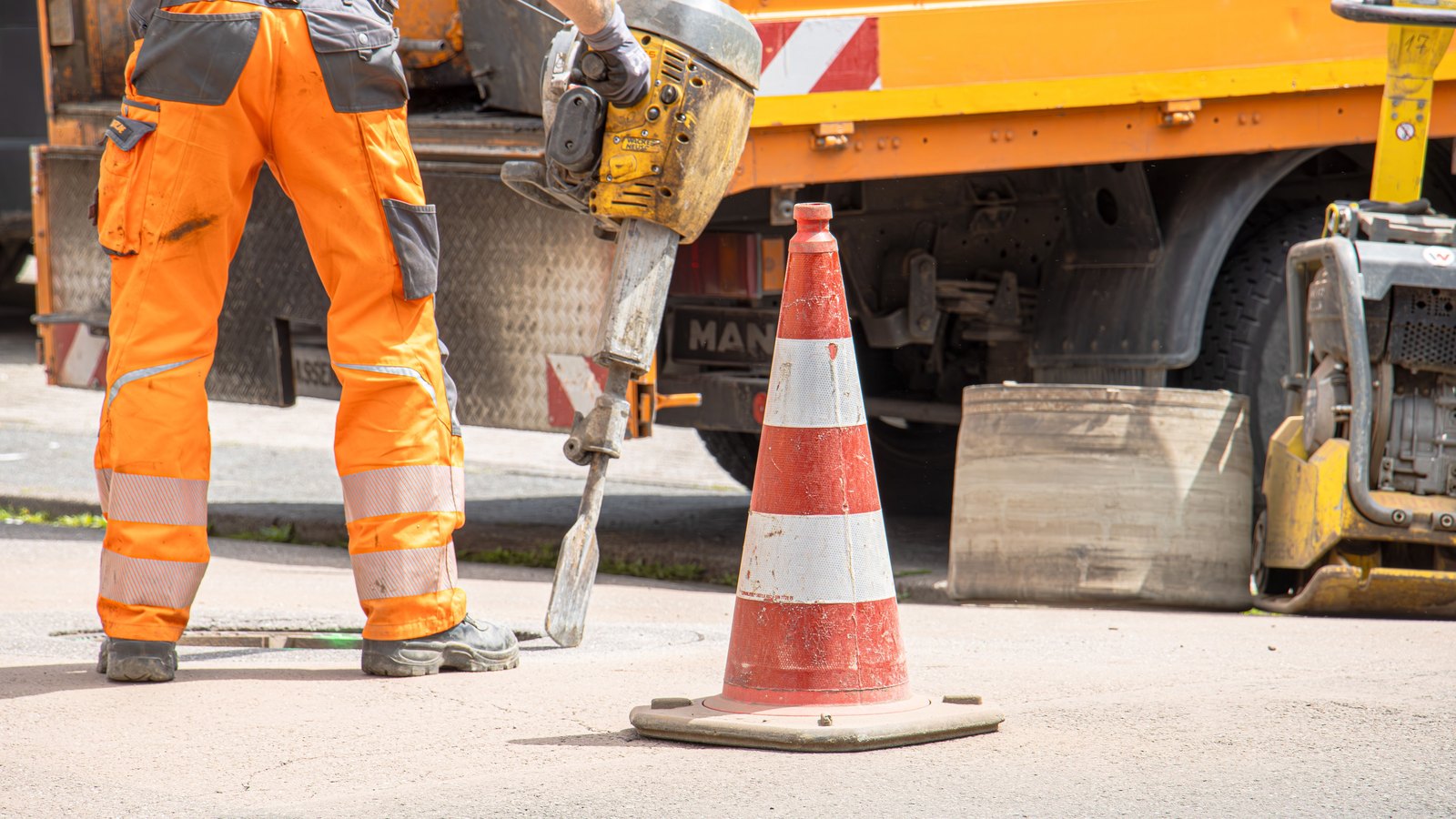 A road worker seen from the waist down in orange high visibility trousers, standing on grey asphalt and holding a jackhammer, with a weathered orange and white traffic cone in the foreground and the side of an orange works truck behind, no face visible and no agency name, number plate or place identifiable anywhere in the frame. A generic illustration, not a photograph of this event.