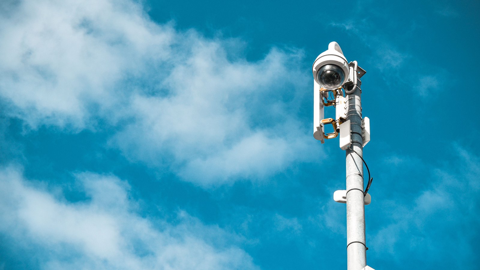 A white dome shaped surveillance camera and its housing mounted near the top of a galvanised metal pole, photographed from below against a blue sky with scattered white cloud. No lettering, marking, landmark or number plate is visible anywhere in the frame.