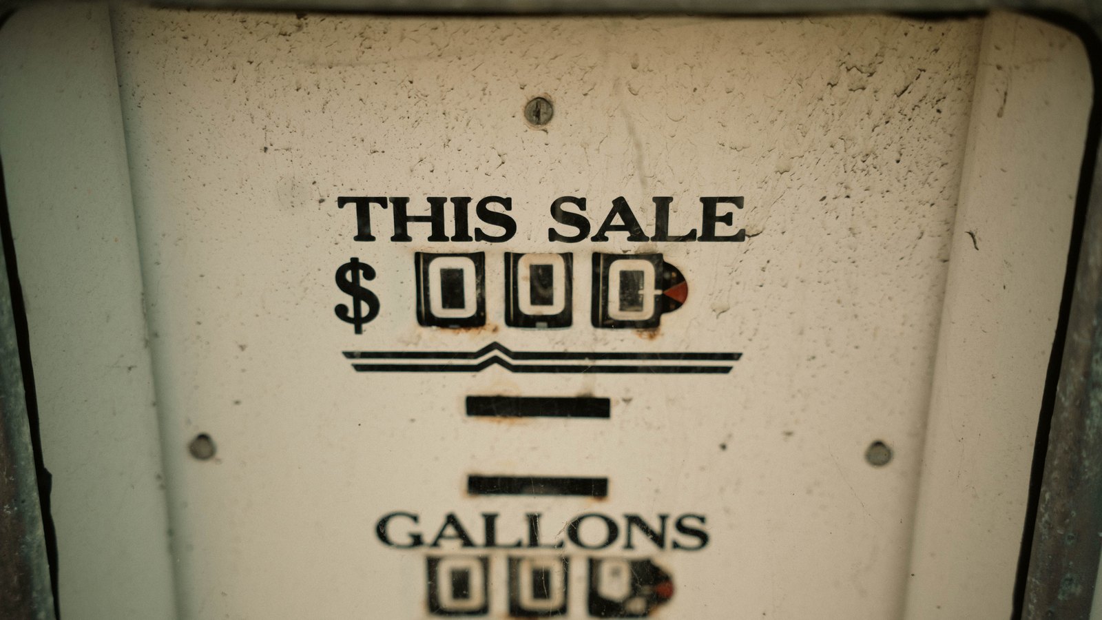 Close-up of the weathered enamel face of an old mechanical gasoline pump, reading THIS SALE in dollars above GALLONS, the numeral wheels all set to zero and rust spotting the panel. No brand name, person, building, road or place is in frame. Stock photo, not a photograph of this event.
