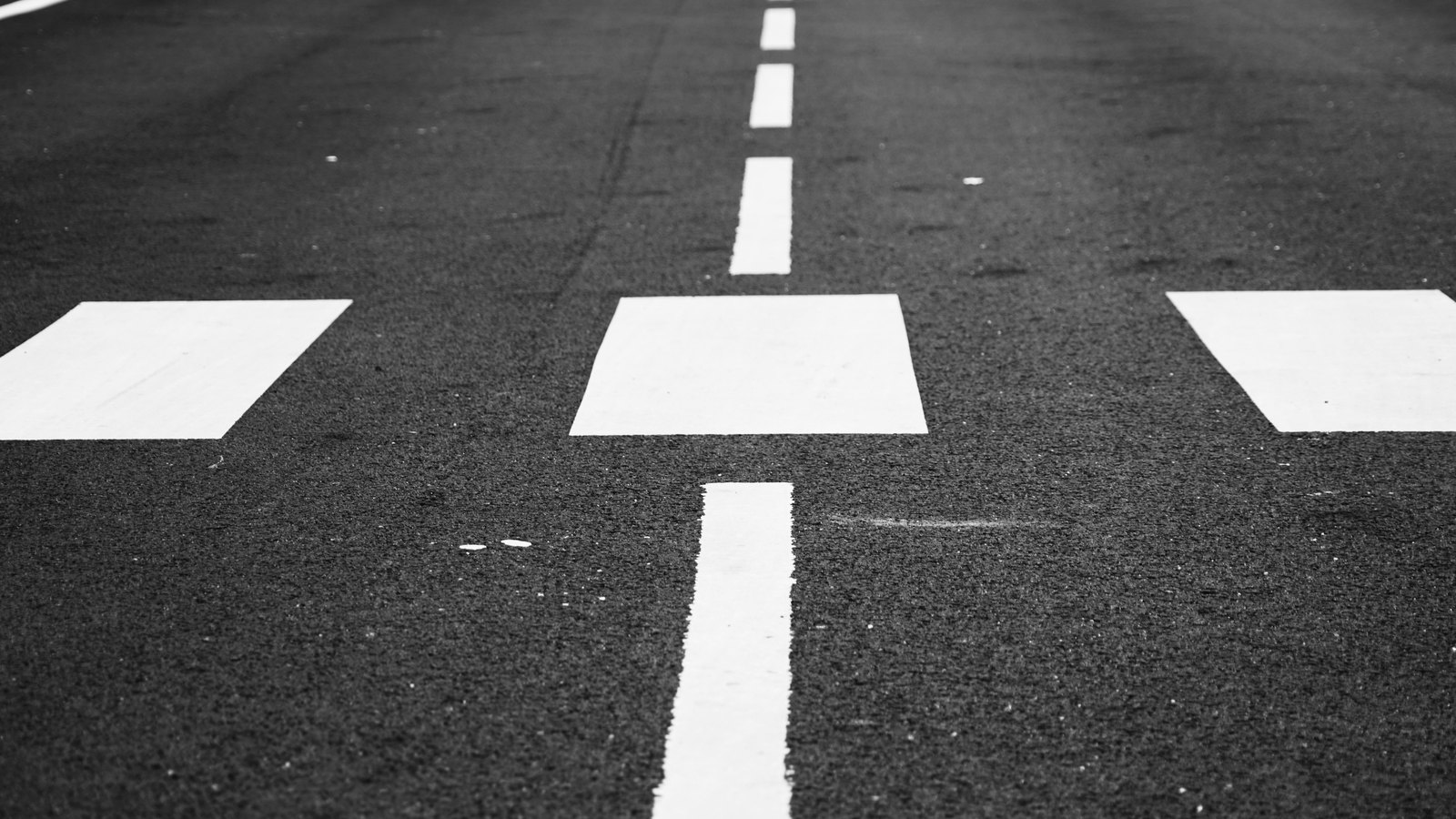 A low black and white view along coarse asphalt, with a broken white lane line running away from the camera between painted white blocks.