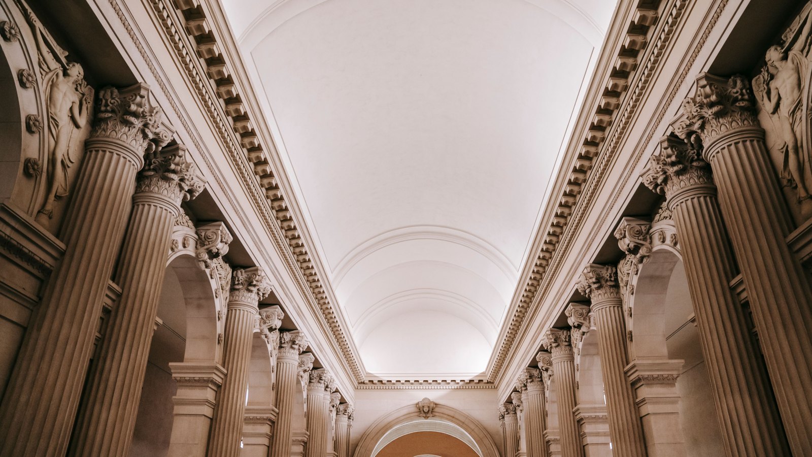 A long colonnade of fluted stone columns with carved capitals running down both sides of a tall hall, under a plain white barrel-vaulted ceiling, with a small arched window of blue and white glass at the far end. An illustrative photograph of a monumental interior, not any building connected with this story.