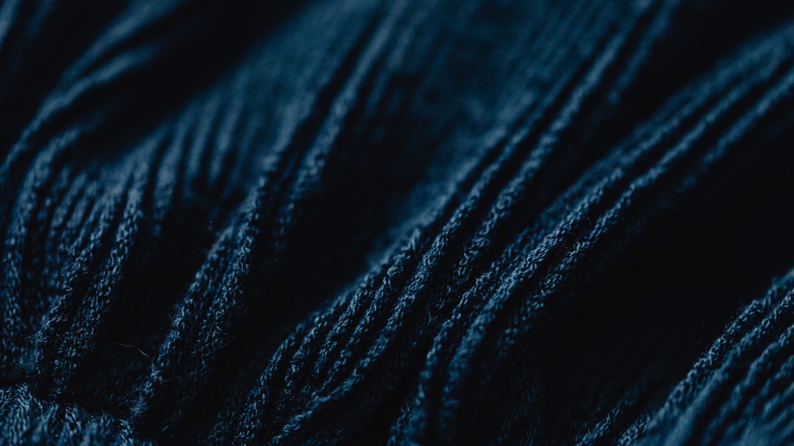 Dark navy knitted fabric photographed close up, its ribbed diagonal folds catching a low raking light against near black shadow.