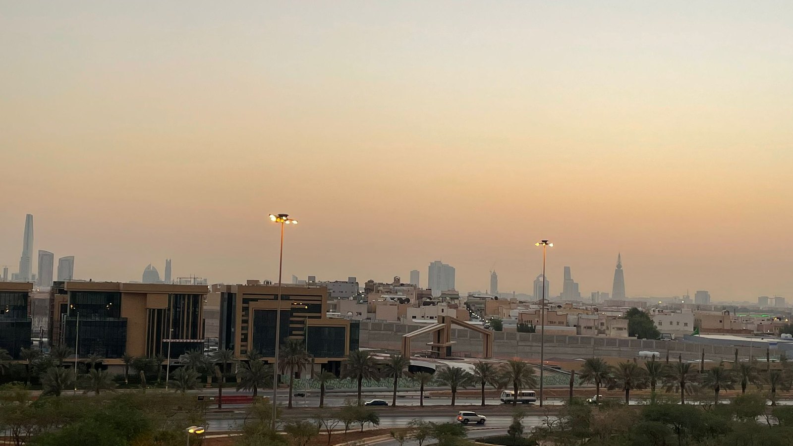 The Riyadh skyline beyond low-rise buildings and palm-lined roads in evening haze.