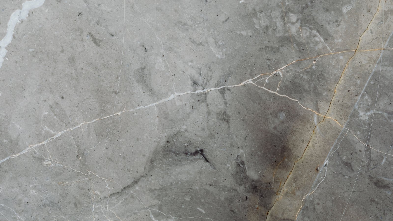 A close photograph of a polished grey stone slab filling the whole frame, pale and dark grey mottling crossed by thin white and ochre veins running diagonally, with faint hairline scratches across the surface. No text, brand mark, person, place or object edge is in view.