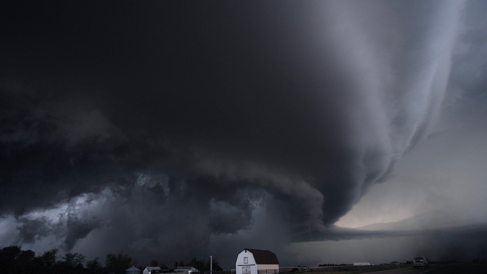 A supercell thunderstorm filling the sky above flat Kansas farmland, with a white barn and grain bins small along the horizon underneath it.